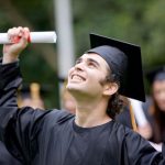 A proud graduate holds his degree and smiles towards the sky
