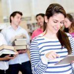 A woman stands with an iPad while three people stand behind her with large books in a library